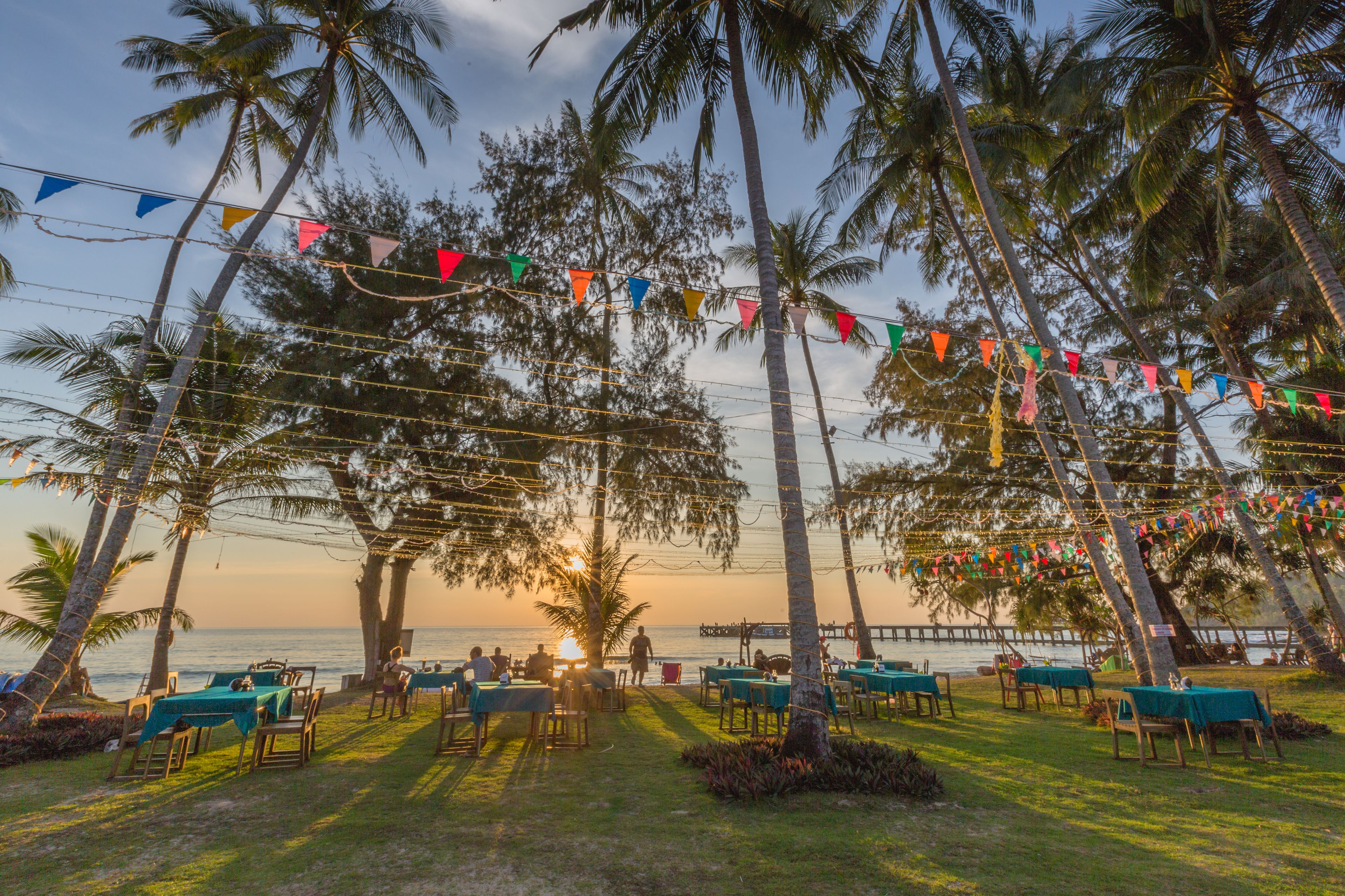 A beachside restaurant with outdoor seating, palm trees, and colorful flags.