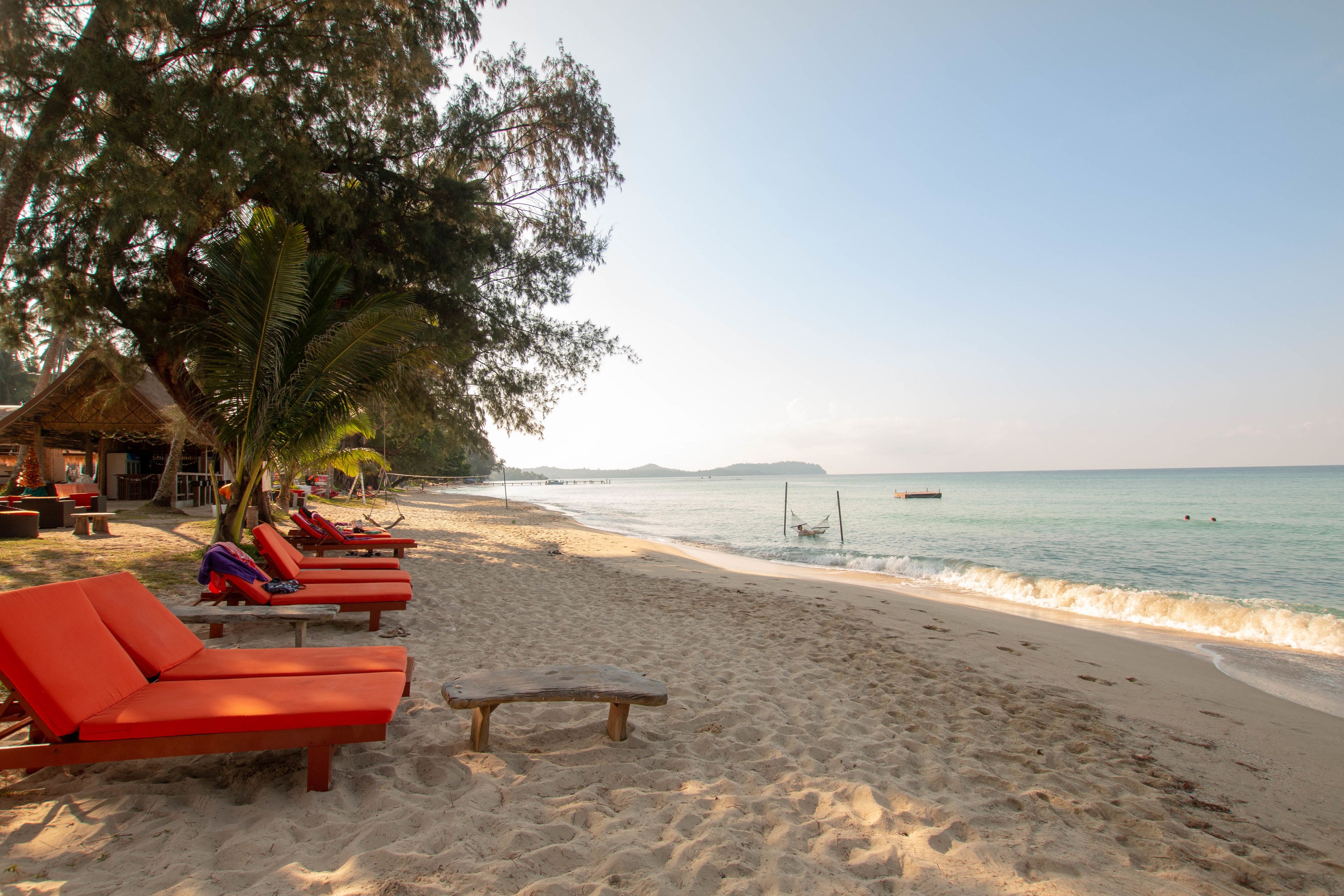 A beach with red lounge chairs and a wooden bench, a thatched-roof hut, and a calm sea with a boat in the distance.