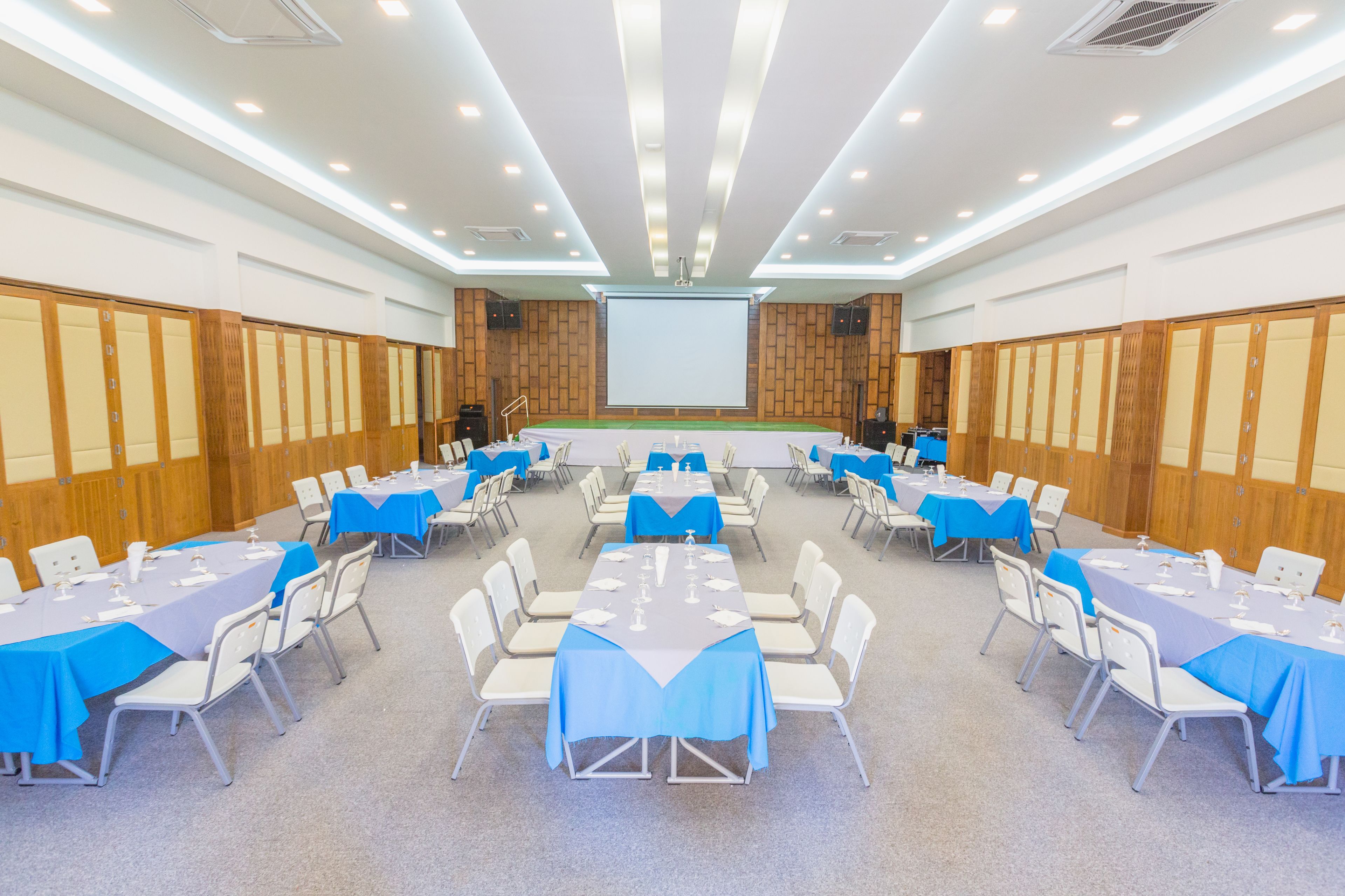 A conference room with multiple tables and chairs, a large screen at the front, and wooden paneling on the walls.