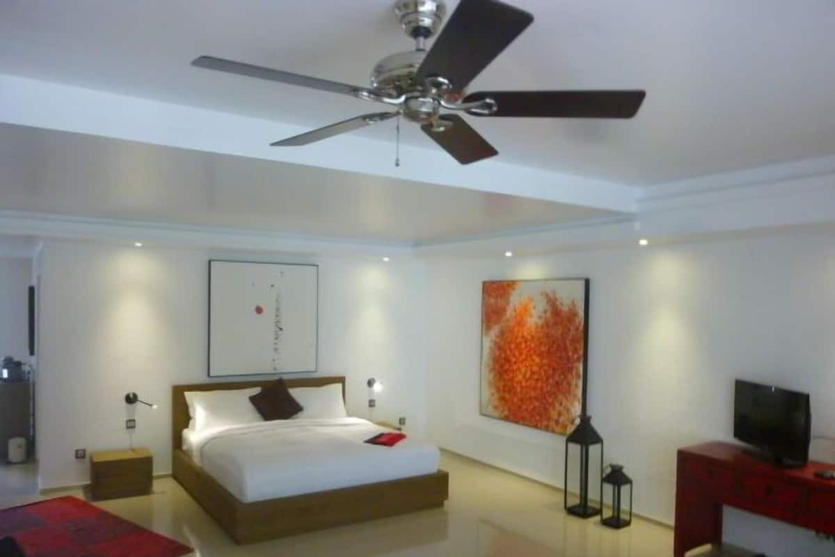 Une chambre moderne avec un lit, un ventilateur de plafond, des œuvres d’art accrochées aux murs, un téléviseur et un bureau rouge.