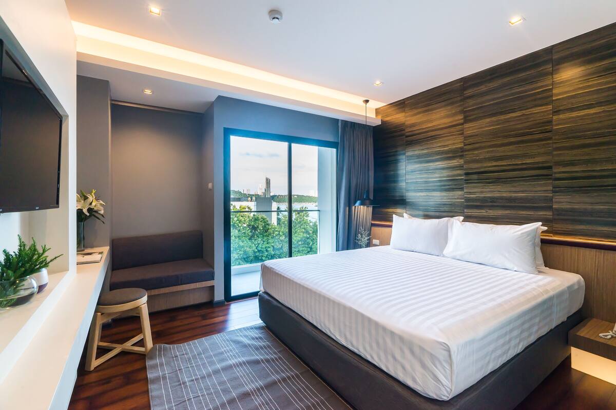 A modern hotel room with a large bed, a desk, and a view of the city through a sliding door.