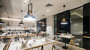 A modern dining area with multiple tables, each set with white tablecloths, plates, and cutlery. There are pendant lights hanging above the tables, and the room has a contemporary design with a mix of natural and artificial lighting. - Sea Me Spring Tree Hotel - Adults only (Pattaya)