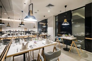 Un comedor moderno con varias mesas, cada una con manteles blancos, platos y cubiertos. Hay lámparas colgantes sobre las mesas, y la habitación tiene un diseño contemporáneo con una combinación de iluminación natural y artificial.