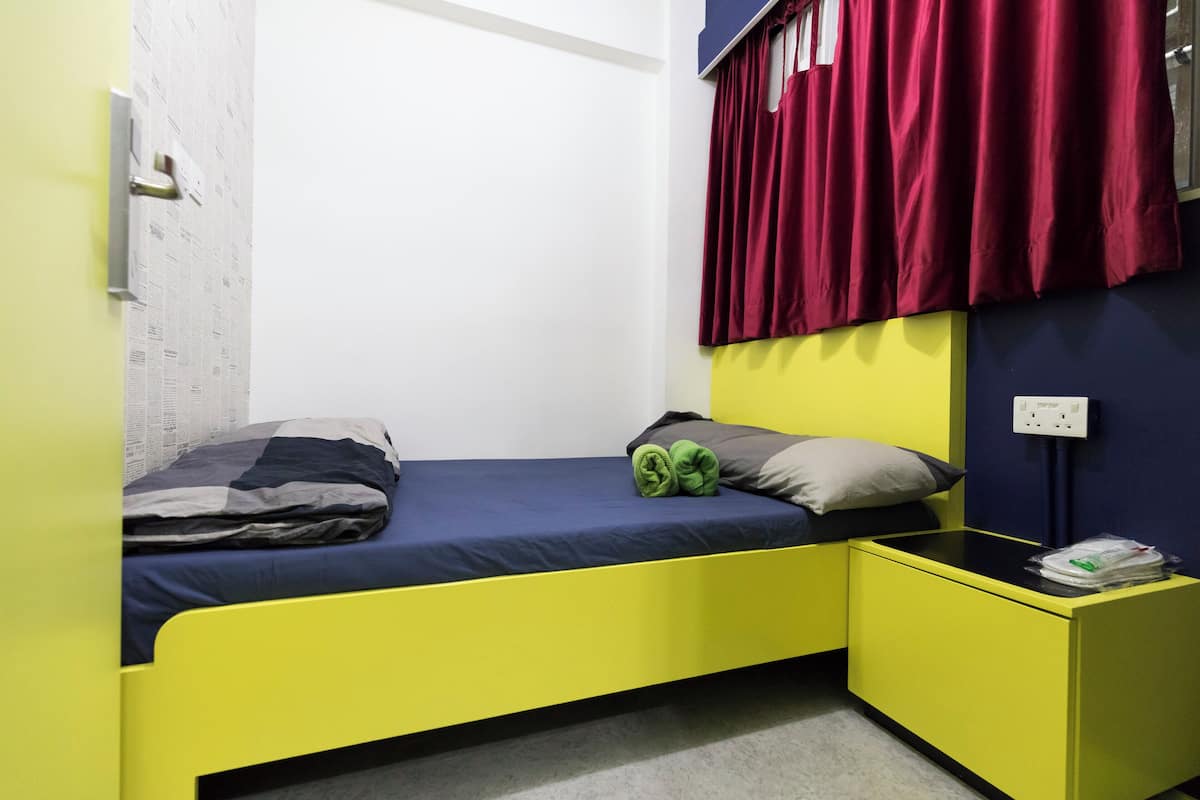 Ein kleiner, hell gefärbter Raum mit einem Einzelbett, einem Nachttisch und einem Fenster mit roten Vorhängen.