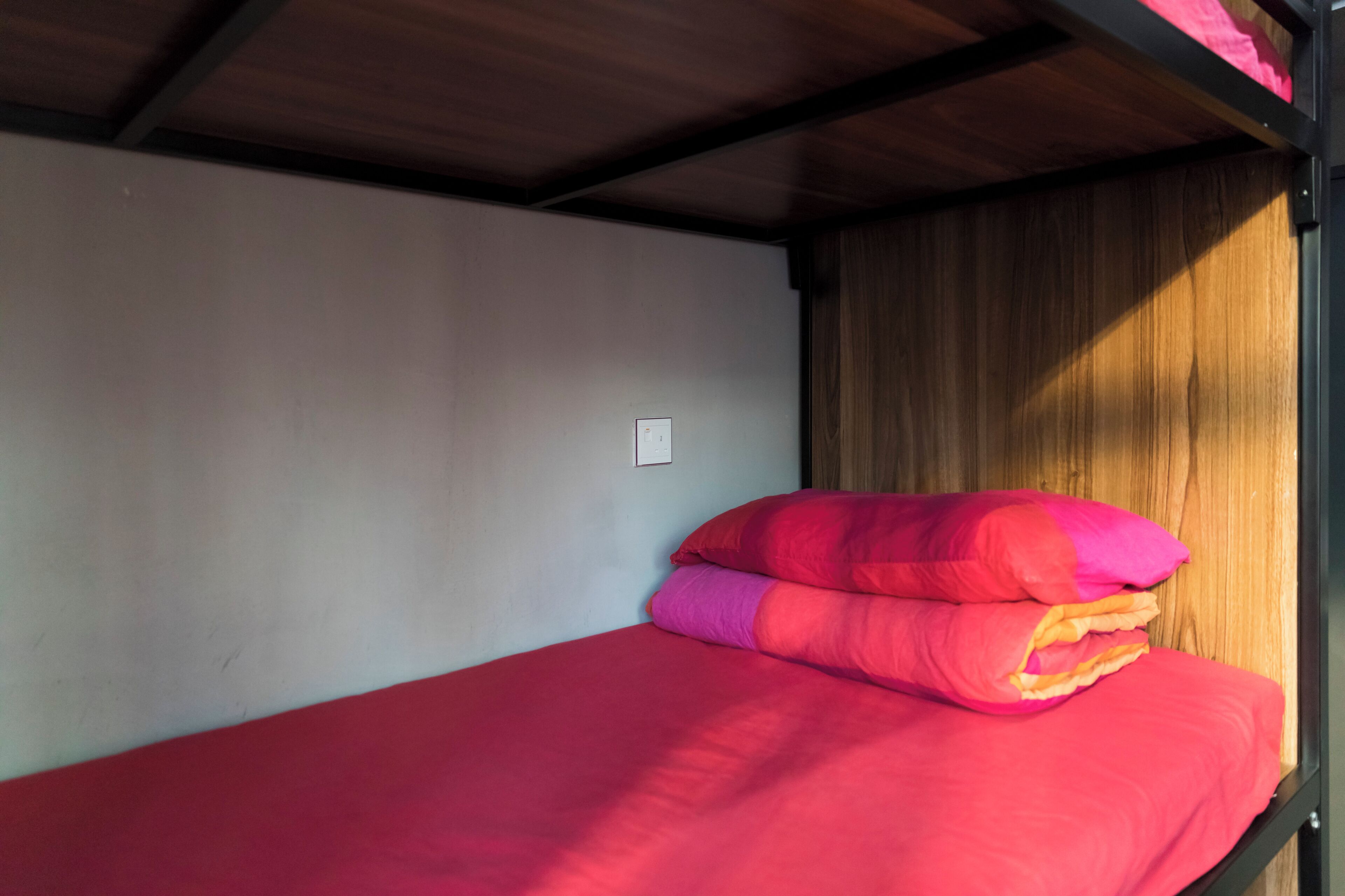 Un lit simple avec un matelas rouge et des oreillers roses, dans une pièce dont le plafond est orné de poutres en bois et un des murs est blanc.