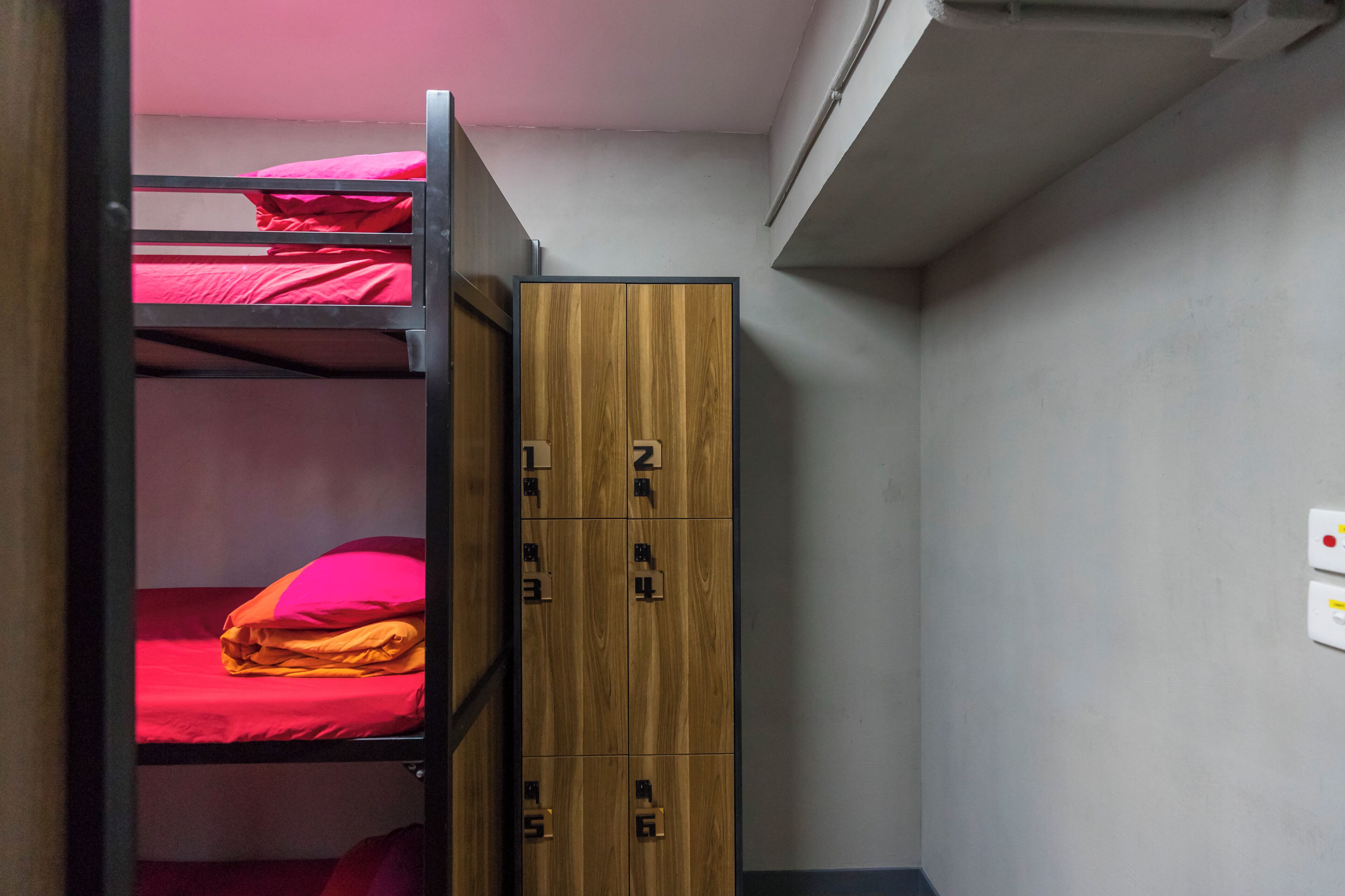 A bunk bed with red bedding, a wooden wardrobe with multiple compartments, and a light switch on the wall.