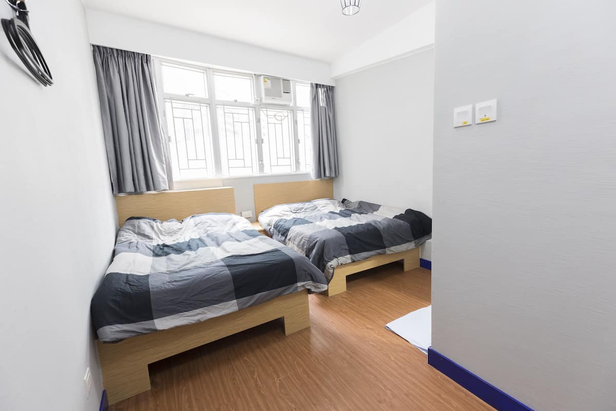Ein Zimmer mit zwei Betten, Holzboden, einem fenster mit Vorhängen und einer weißen Wand mit zwei Lichtschaltern.