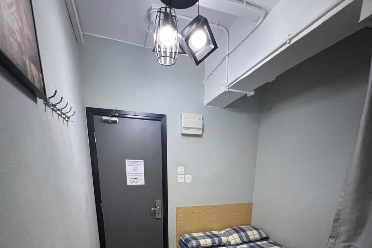 Ein kleines Zimmer mit einem Einzelbett, Holzboden, einem Bett mit kariertem Überwurf, einer Deckenleuchte und einer Tür mit einem Schild.