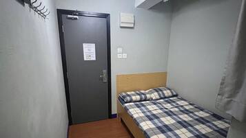 A small, single-bed room with a wooden floor, a bed with a plaid bedspread, a ceiling light fixture, and a door with a sign.