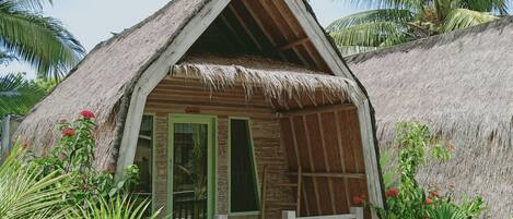 Un bungalow au toit de chaume, doté d’un porche en bois, entouré d’une végétation tropicale et bordé d’un sentier en pierre.