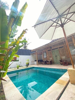A swimming pool with a tiled edge, surrounded by a wooden deck and a covered seating area with chairs and a table. - Villa Coco Republic (Gili Air)