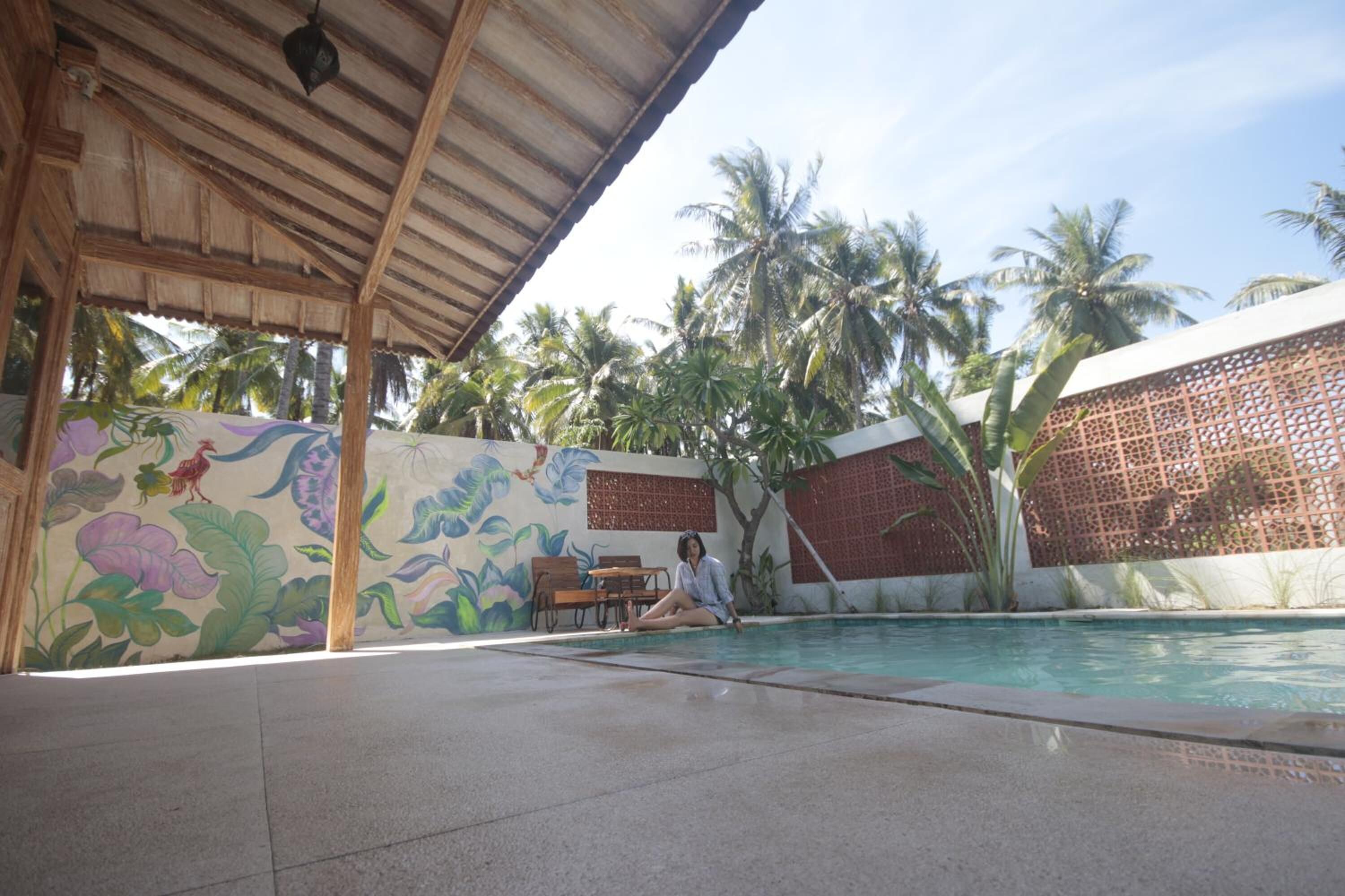 Un espace piscine doté d’un coin salon abrité, de palmiers et d’une fresque murale.