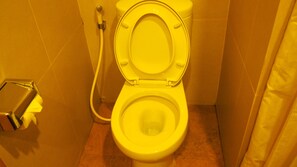 Des toilettes jaunes, équipées d’un siège-bidet, dans une petite salle de bain aux murs carrelés et dotée d’un rideau.