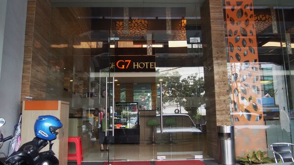 The entrance of G7 Hotel with a glass facade, a visible interior with a reception desk, and a motorcycle parked outside.