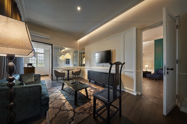 A modern living room with a sofa, coffee table, and TV. There is a dining area with chairs and a table. The room has a large window and a door leading to another room.