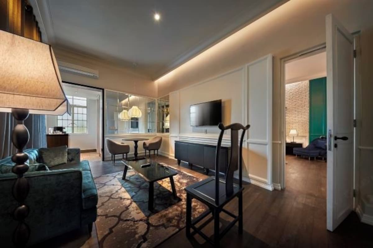 Un salon moderne avec un canapé, une table basse et des fauteuils, un téléviseur à écran plat fixé au mur, et un coin cuisine visible à travers une porte ouverte.