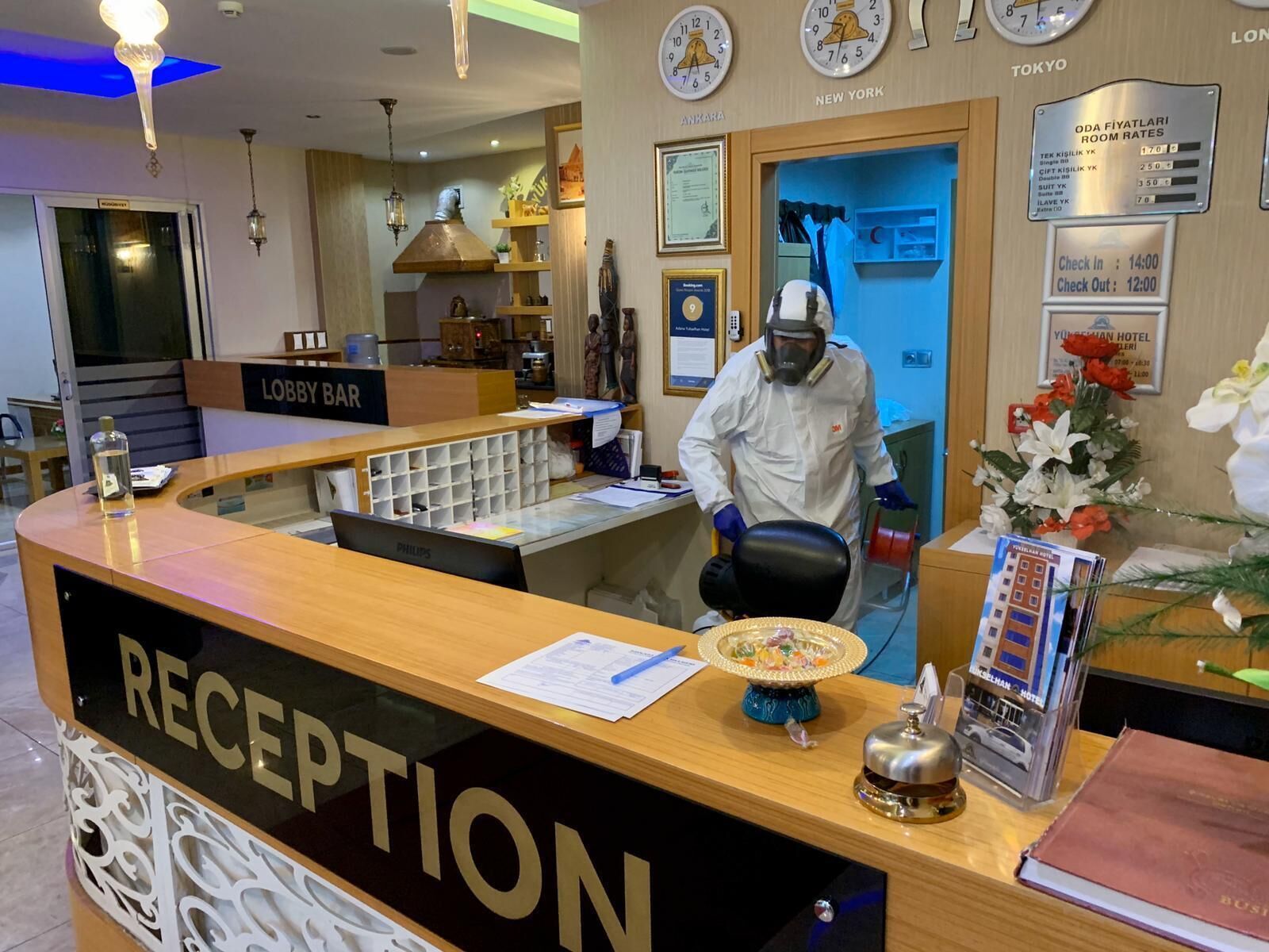 Un espace d’accueil d’hôtel comprenant un comptoir de réception portant l’inscription « RÉCEPTION », un bar dans le hall et une horloge affichant les tarifs des chambres à Tokyo, New York et Londres.