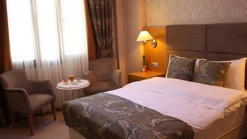A hotel room with a large bed, two armchairs, a small round table with breakfast items, a nightstand with a lamp, and a view of the outdoors through sheer curtains.