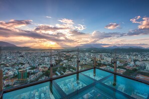 Vista panorámica desde un edificio de gran altura con piso de vidrio, que domina un paisaje urbano con diversos edificios y montañas distantes.