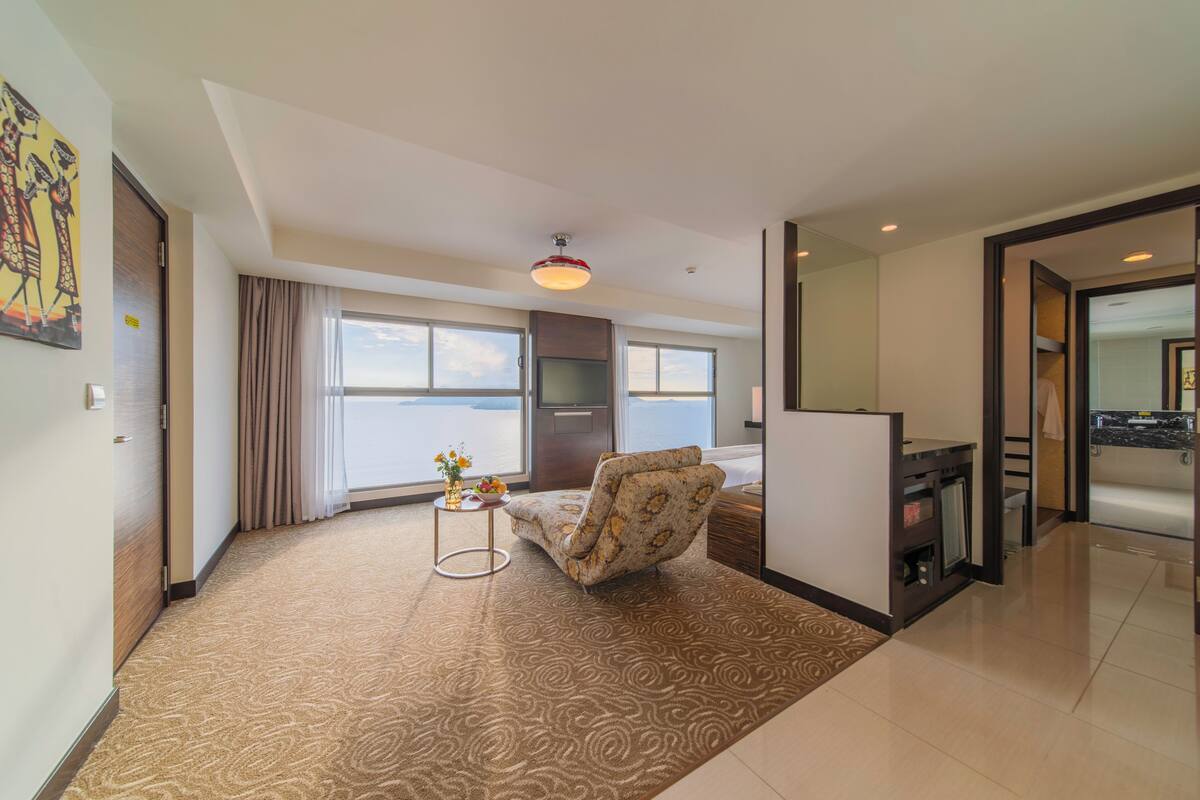 A hotel room with a large window, a sofa, a small table with a vase, and a bathroom visible through an open door.