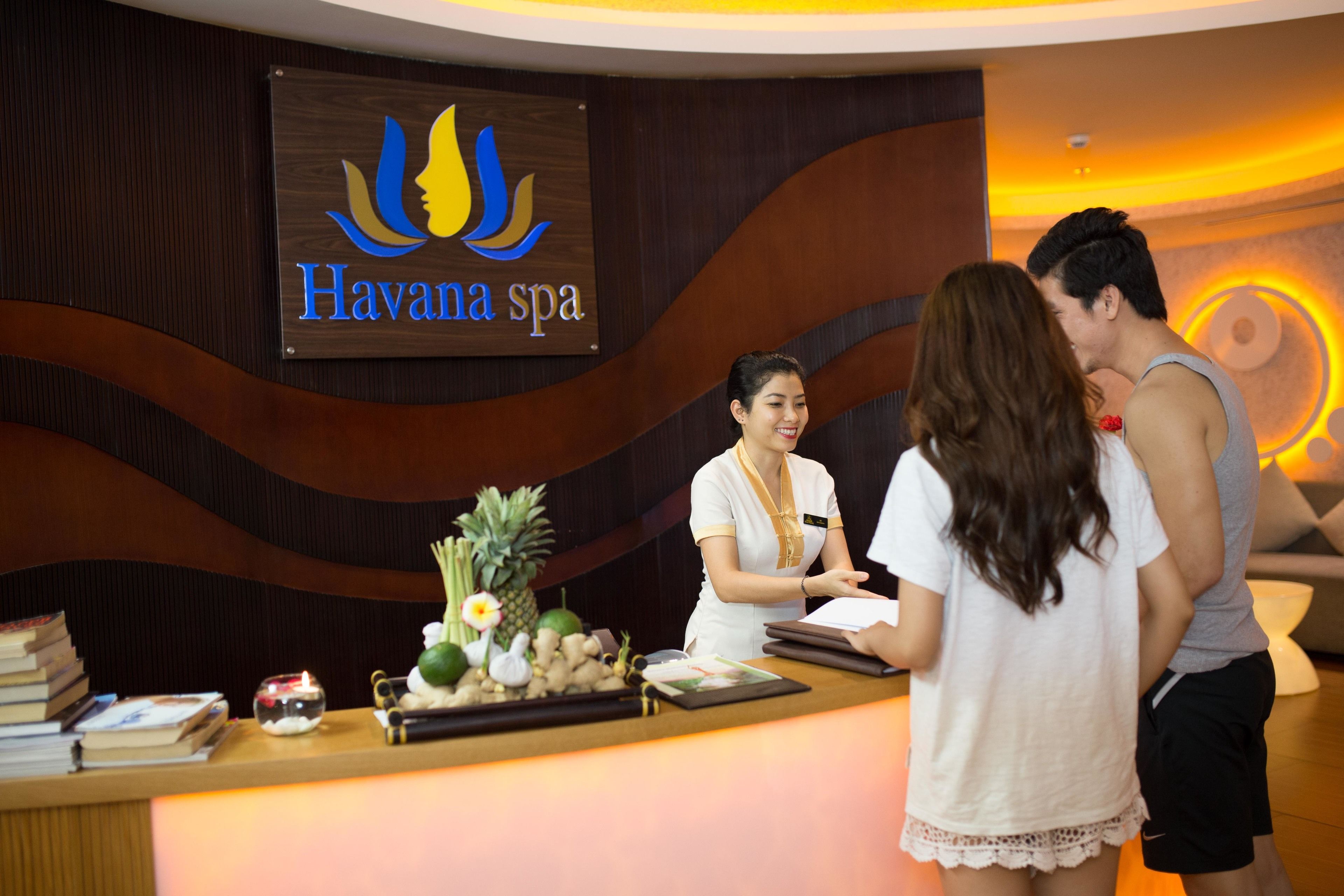 A reception desk at Havana spa with a wooden sign, a receptionist, and guests.