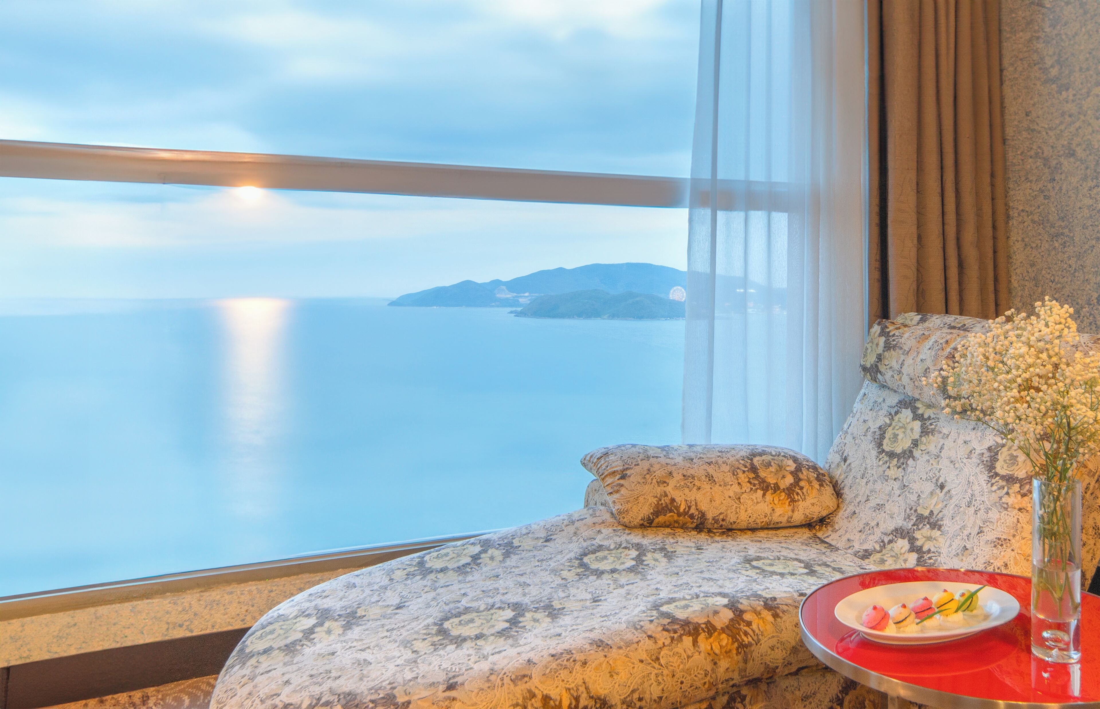 A room with a view of the sea, a sofa with a floral pattern, and a table with a plate of food and a vase of flowers.