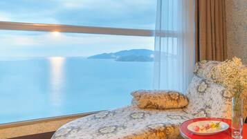 A room with a view of the sea, a sofa with a floral pattern, and a table with a plate of food and a vase of flowers.