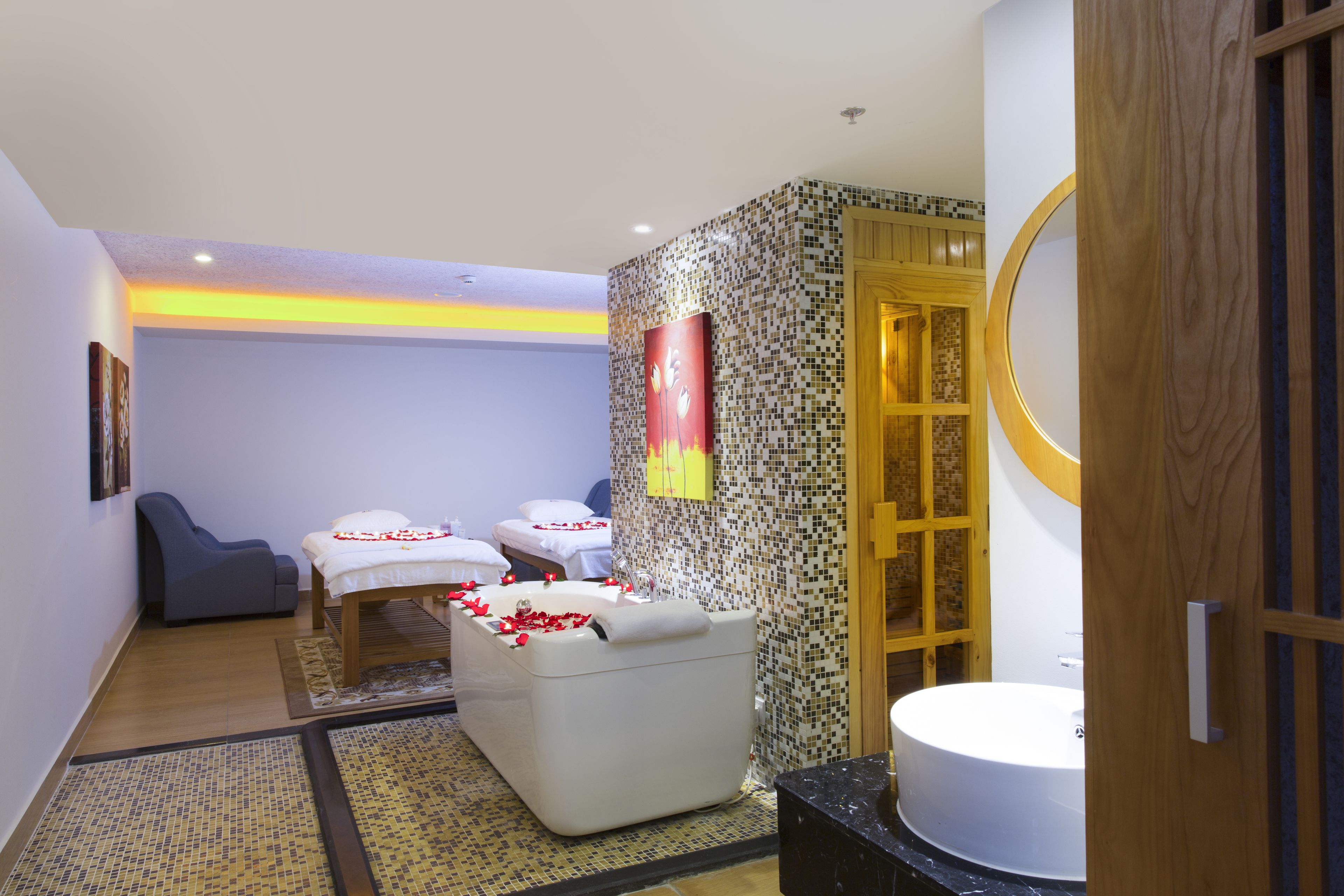A modern spa room with a massage table, a massage chair, and a bathtub with rose petals.