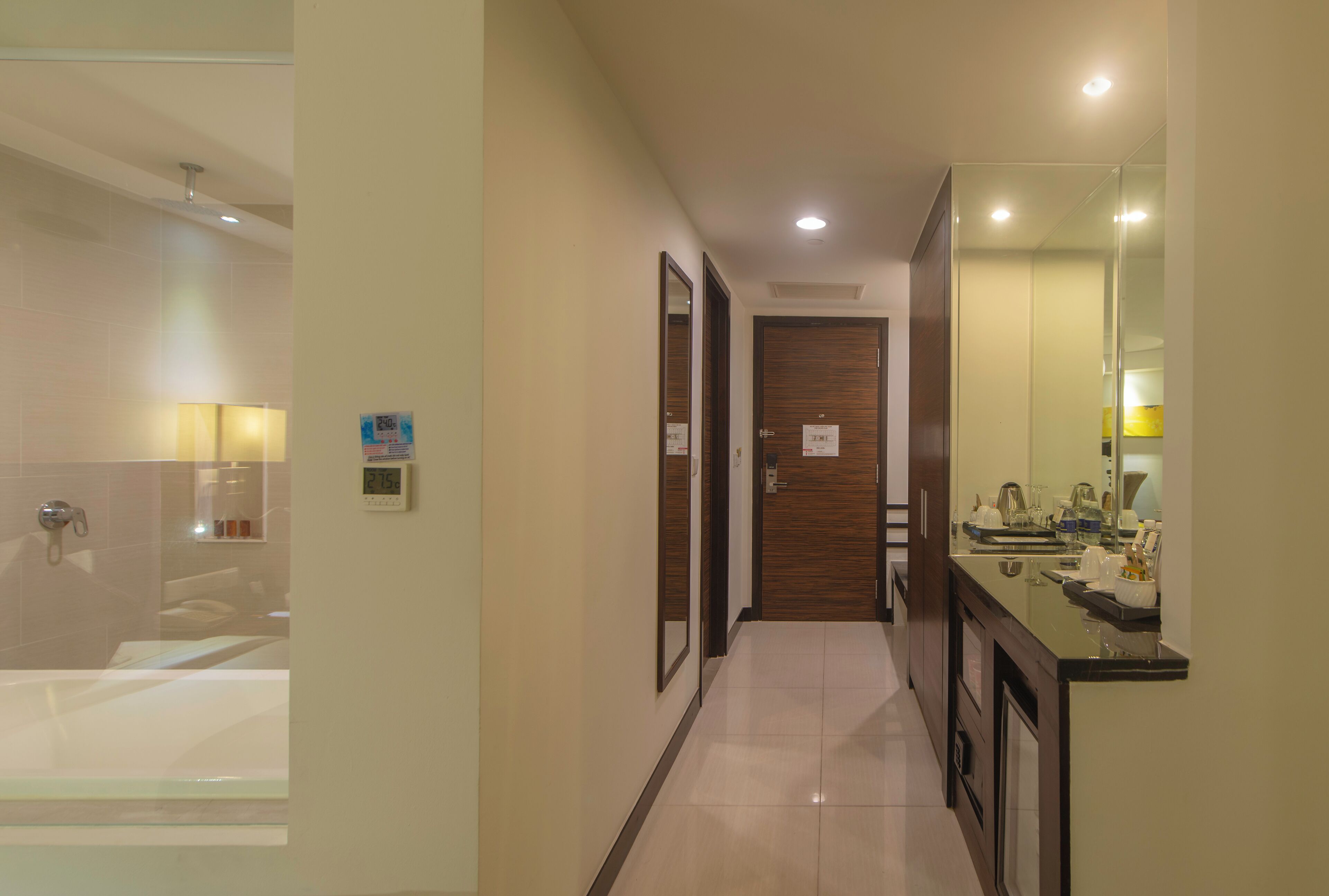 A hotel hallway with a bathroom on the left, a wooden door in the center, and a bathroom with a sink and mirror on the right.
