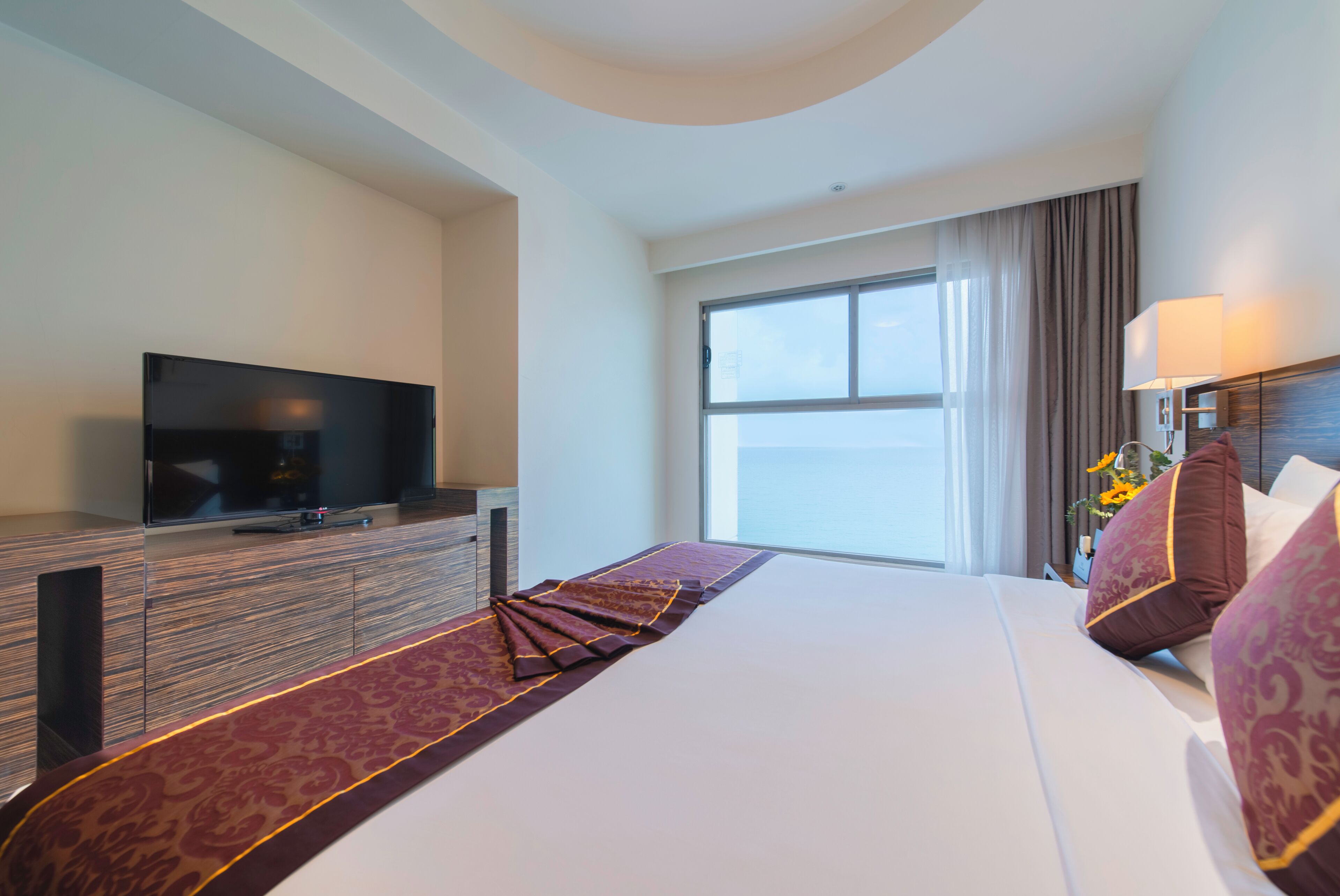 Een hotelkamer met een groot bed, een flatscreen televisie op een houten dressoir, een groot raam met gordijnen en een nachtkastje met een lamp.