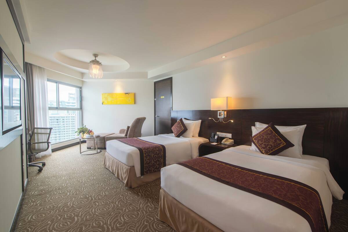 A hotel room with two beds, a desk, a chair, a window with curtains, and a ceiling light.