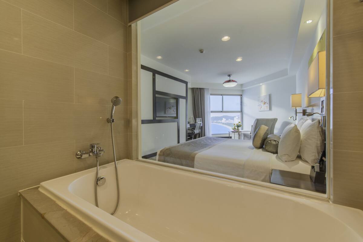 A modern hotel room with a large bathtub, a bed with pillows, a television, and a view of the city.