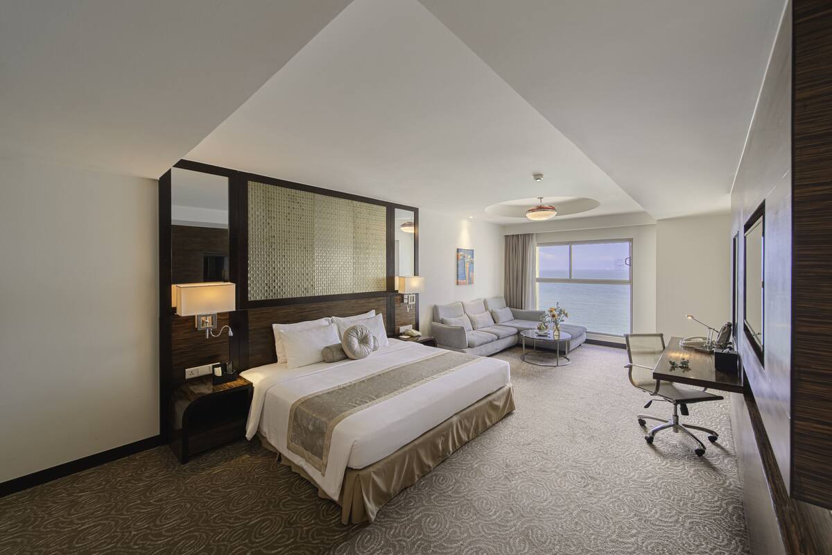 A modern hotel room with a large bed, a seating area with a sofa and armchairs, a desk, and a large window offering a view of the sea.