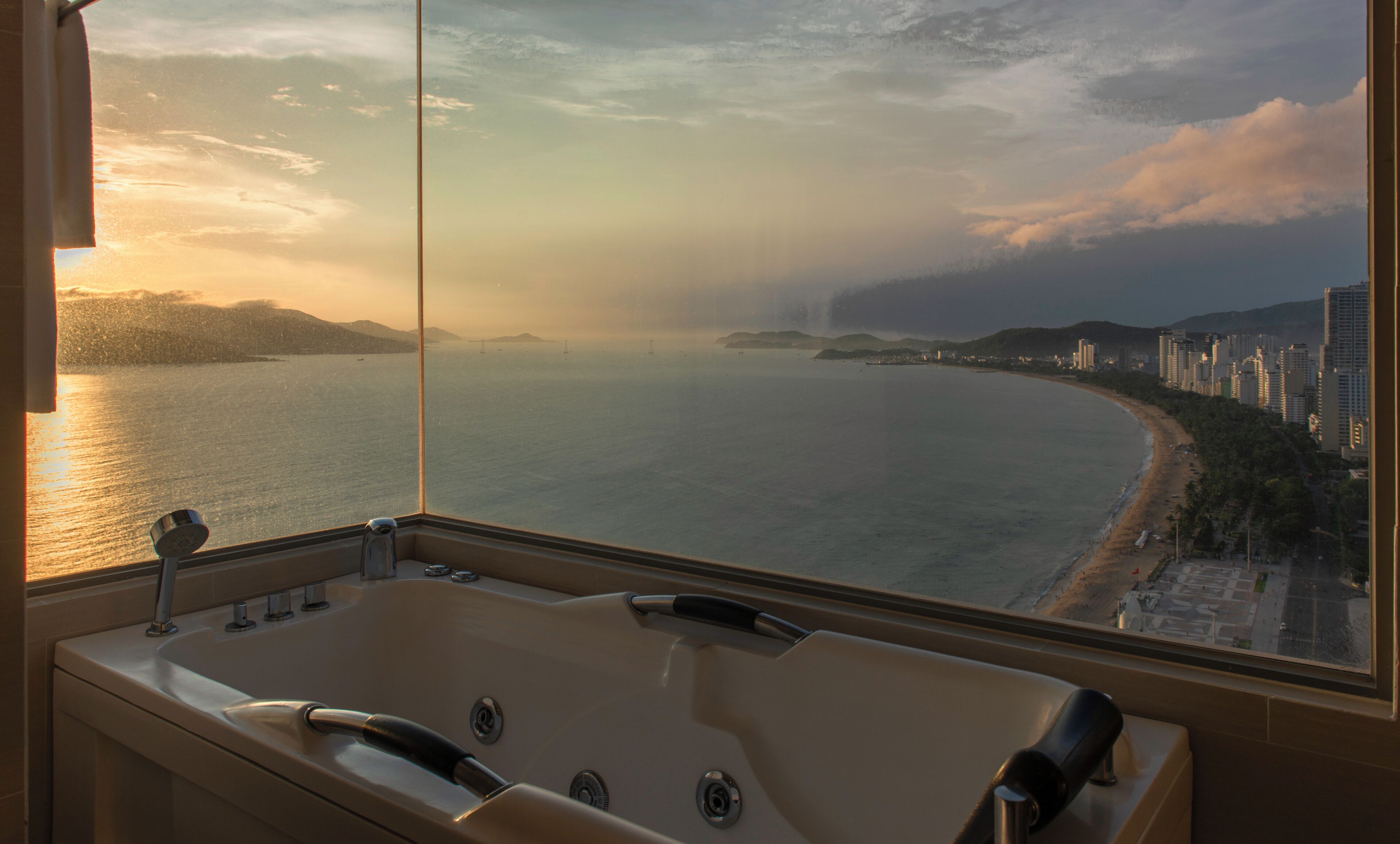 A bathroom with a large bathtub and a view of a coastal cityscape at sunset.