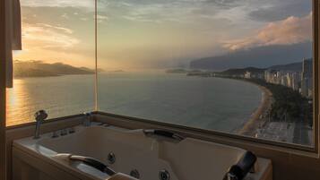 A bathroom with a large bathtub and a view of a coastal cityscape at sunset.