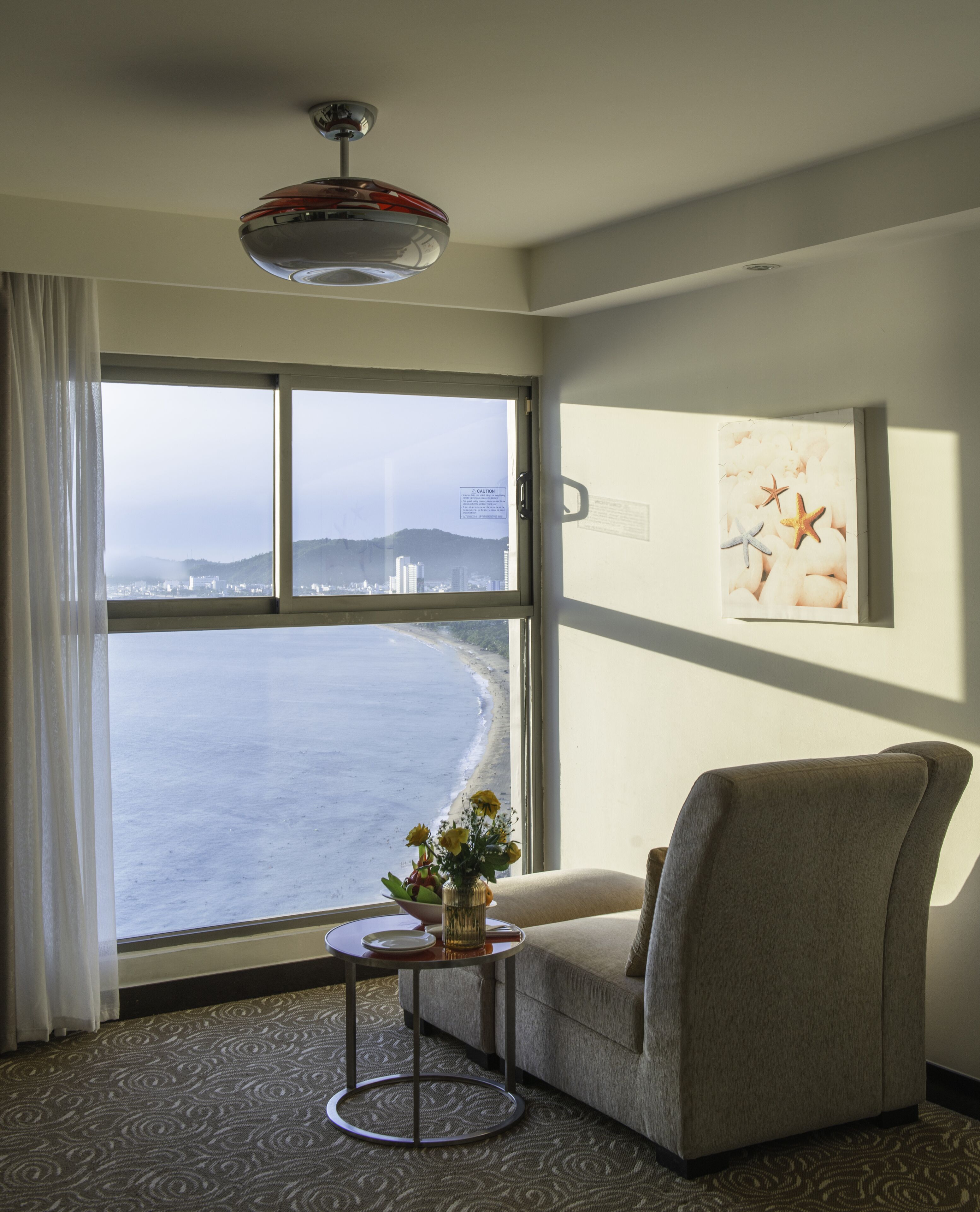 A room with a large window overlooking a beach, a chair, a small table with a vase of flowers, and a ceiling light fixture.