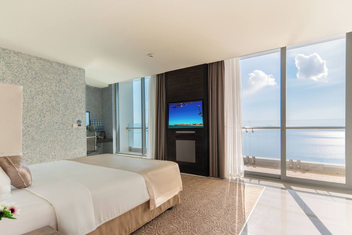 A hotel room with a large bed, a TV, a bathroom, and a balcony with a view of the sea.