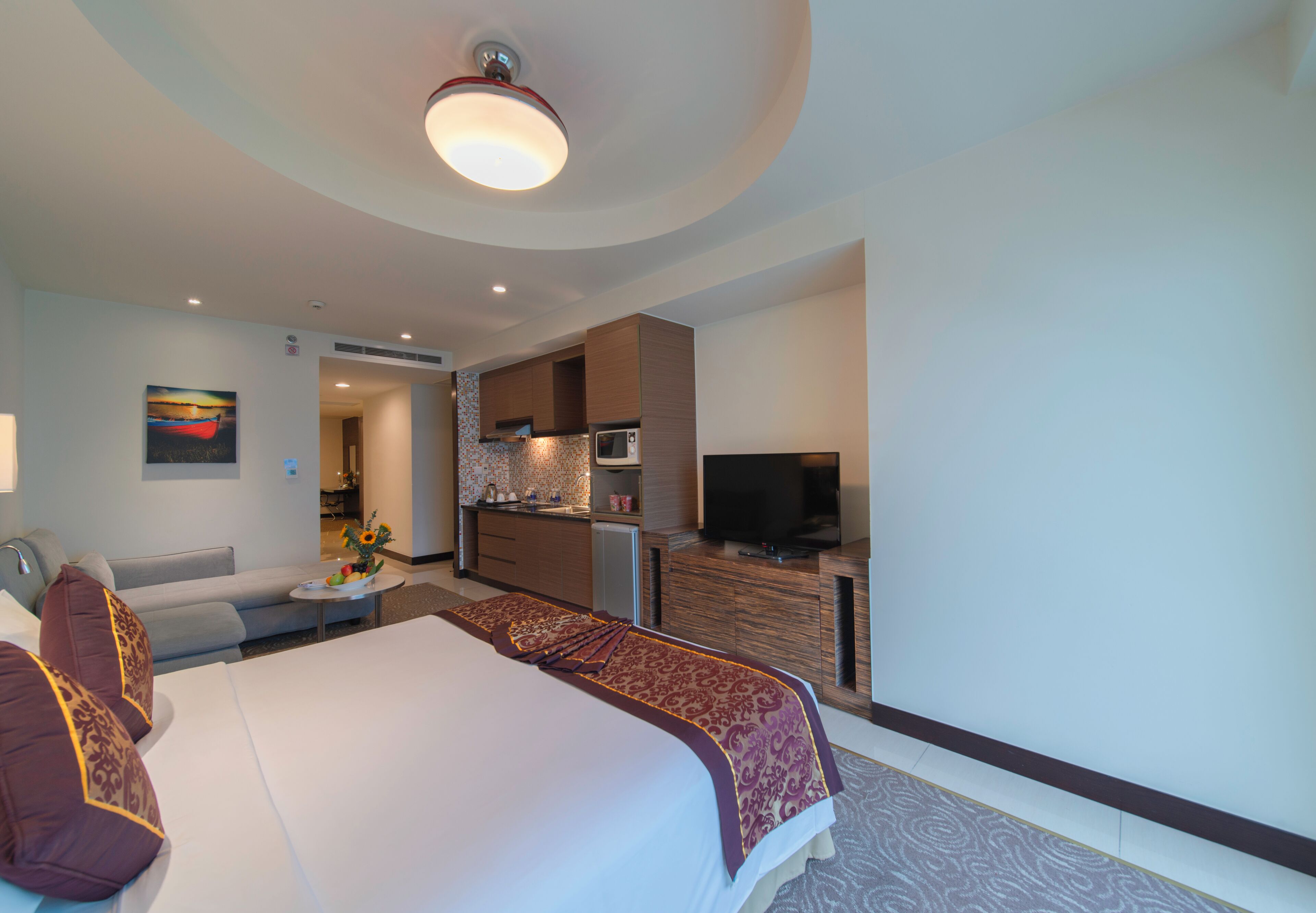 A hotel room with a large bed, a TV, a kitchenette, and a bathroom visible through an open door.