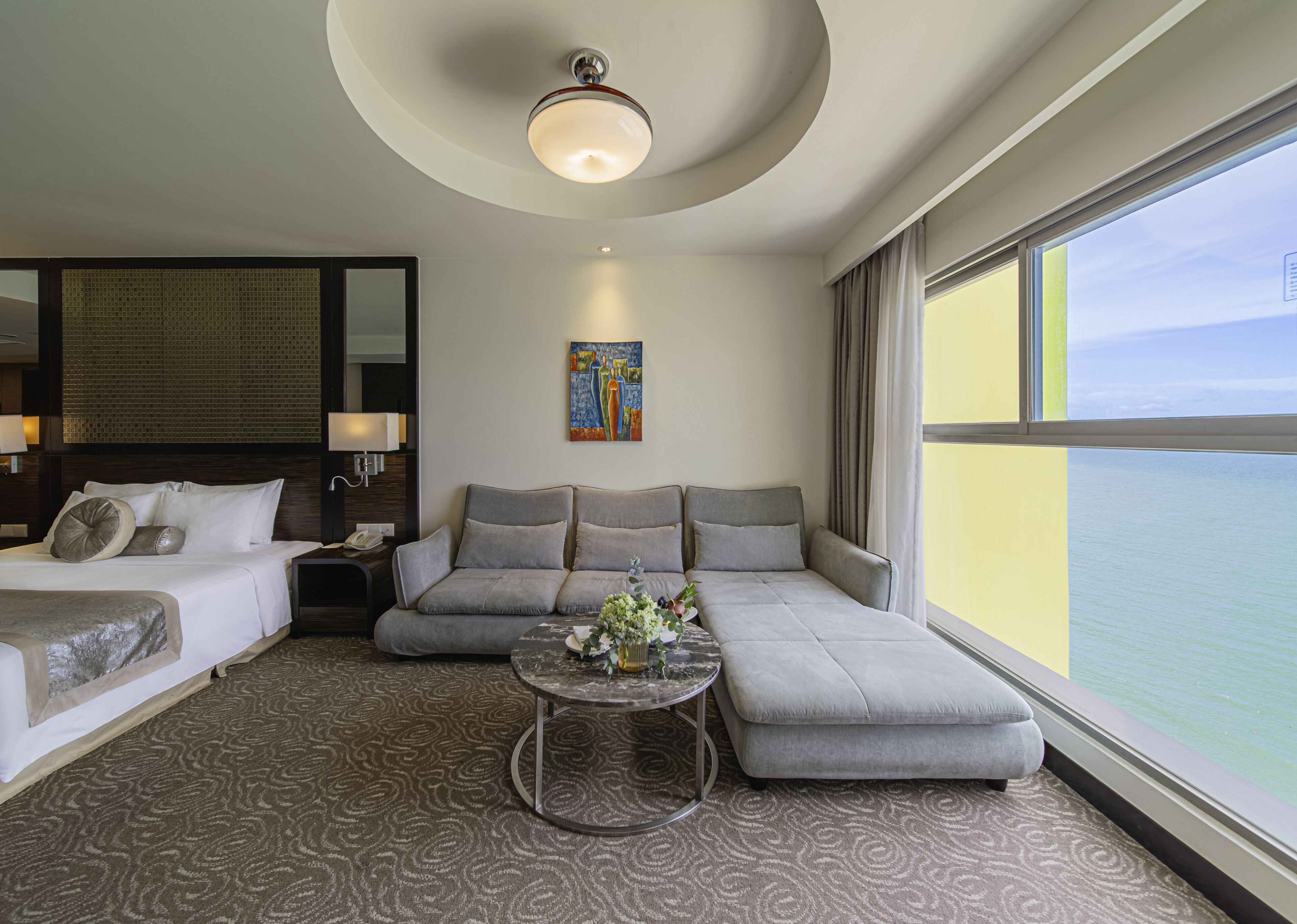 A hotel room with a bed, sofa, and a small table with flowers. There is a painting on the wall and a window with a view of the sea.