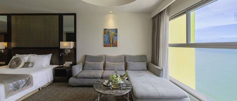 A hotel room with a bed, sofa, and a small table with flowers. There is a painting on the wall and a window with a view of the sea.