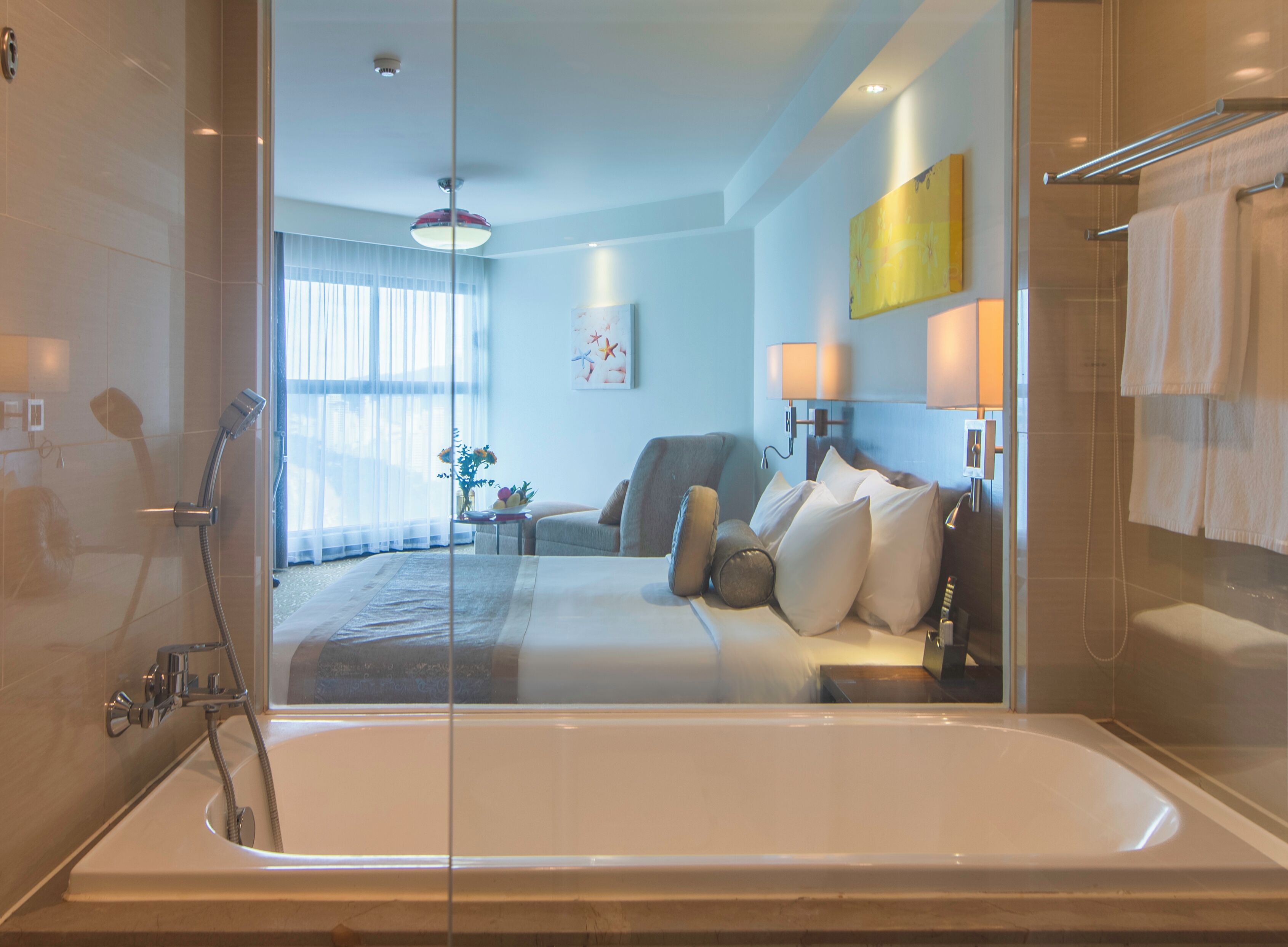 Une chambre d’hôtel moderne dotée d’une baignoire entourée de parois en verre, d’un coin salon confortable avec un canapé et des fauteuils, et d’une grande fenêtre laissant entrer la lumière naturelle.