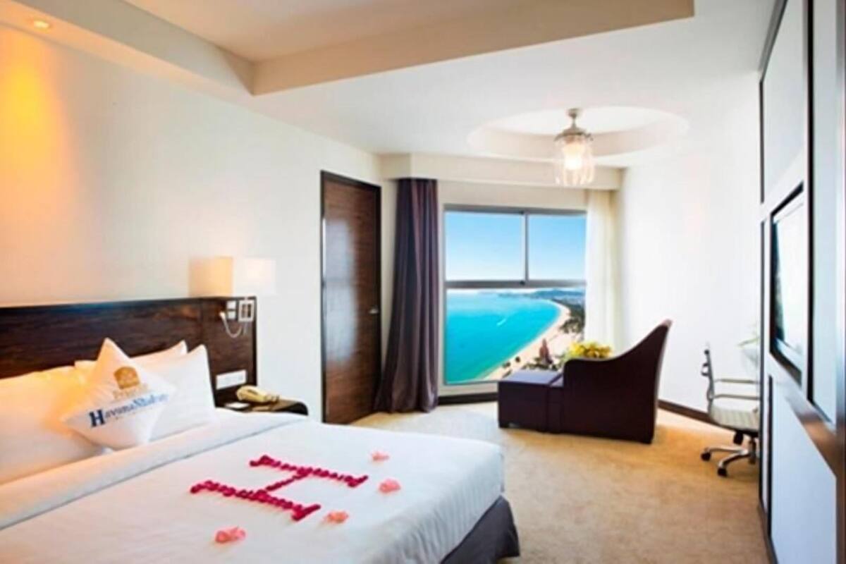 A hotel room with a bed, a desk, a chair, and a large window offering a view of the ocean.