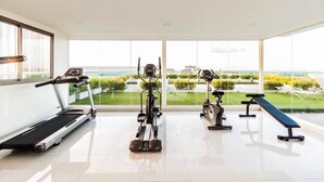 A modern gym with a treadmill, elliptical, stationary bike, and bench. It features large windows with a view of a landscaped area and buildings. - Club Royal Wongamat by PSR ASIA (Pattaya)