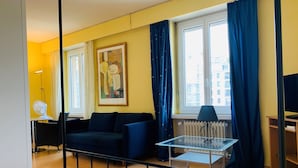 Habitación de hotel con cama, sofá, mesa con lámpara, radiador y ventana con cortinas.