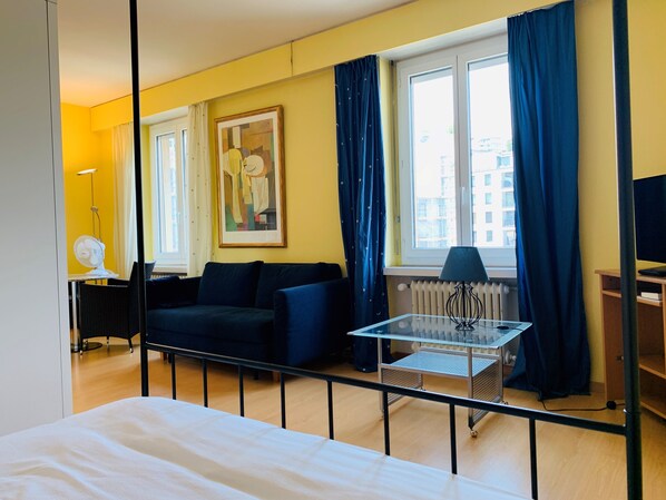 A hotel room with a bed, a sofa, a table with a lamp, a radiator, and a window with curtains.