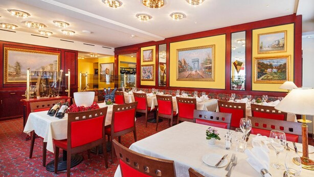 Un restaurante con varias mesas dispuestas para comer, sillas tapizadas de rojo y cuadros en las paredes.