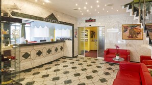 Zona de recepción de un hotel con mostrador, un reloj, una escalera y un sofá rojo.