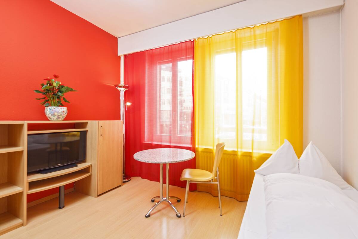 A hotel room with a bed, a TV on a wooden stand, a small table, a chair, and a window with red and yellow curtains.