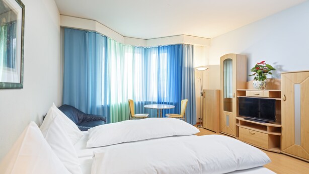 Une chambre d’hôtel avec un lit, une télévision, une petite table, deux chaises et une plante.