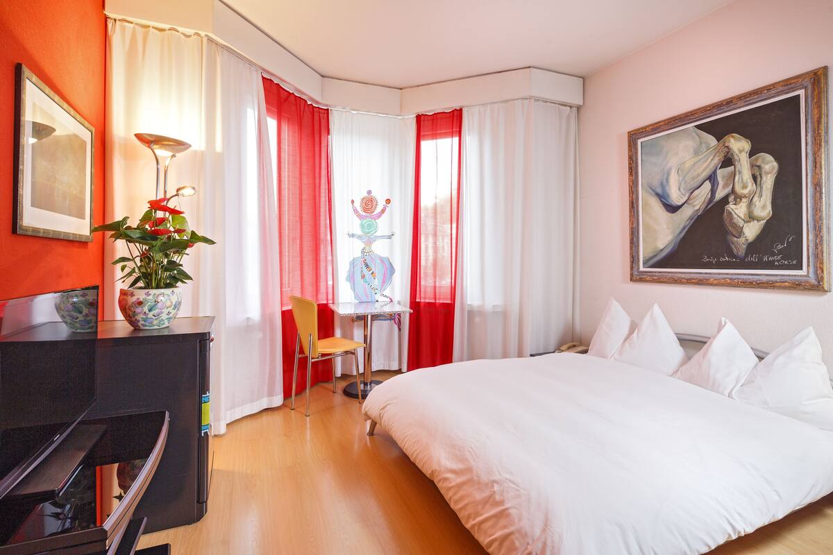 A hotel room with a bed, a desk, a chair, a potted plant, and a painting on the wall.